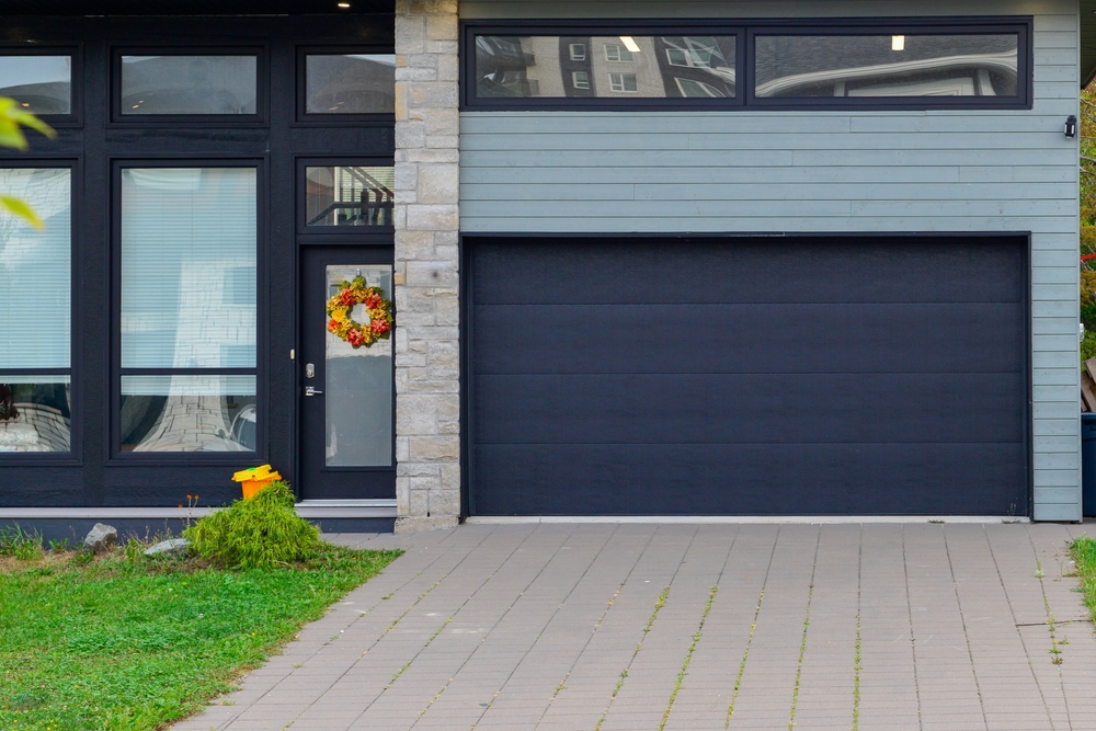 A modern black panel metal exterior garage door. The elegant door is on a luxury dark grey and beige brick contemporary house with a concrete driveway. The house entrance door is glass with black trim